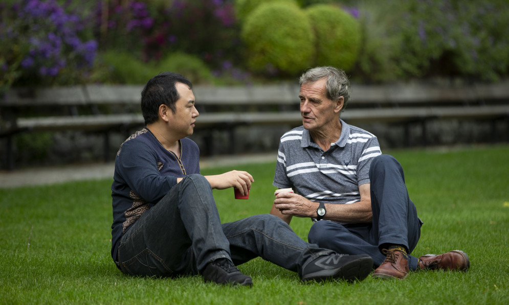 Two men sitting on the grass in a park with coffee cups, talking. 