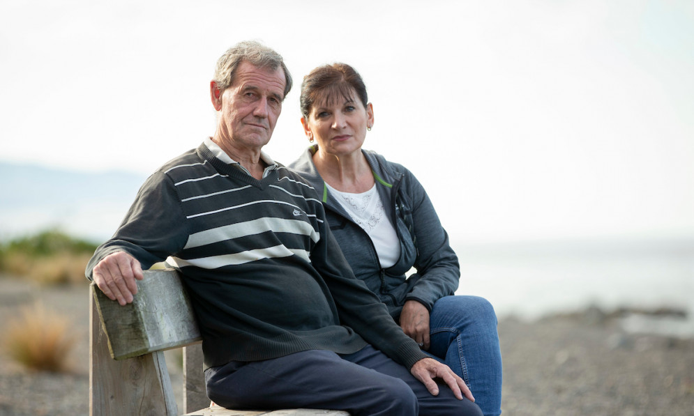 An older man and woman sitting on a bench by the beach, looking at the camera. 